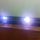Černý lineární LED panel s řadou kulatých světelných zdrojů, z nichž několik intenzivně svítí bíle. Na předním panelu je digitální displej s červeným…