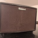 Černý kompaktní kytarový kombo zesilovač s hnědou texturovanou síťovkou. V horní části je umístěno bílé logo Roland. Na hranách šasi jsou viditelné d…