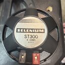 Černý reproduktor s kovovou ochrannou mřížkou. Na středovém bílém štítku je uveden nápis SELENIUM, model ST300, specifikace 8 OHMS a datum 28/03/01.…