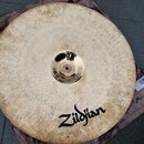 Kruhový mosazný činel s viditelnou Oxidací a patinou po celém povrchu. V horní části je černý nápis Zildjian. Činel leží na hnědé látkové podložce.