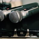 Dvě bezdrátové mikrofony s kovovými mřížkami a černými rukojetmi leží na černém zařízení. Na přední straně zařízení je viditelné logo LD. Mikrofony j…