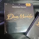 Fotografie zobrazuje dvě uzavřená balení strun. Na čelní straně dominantního balení je zlatým písmem nápis Dean Markley a nad ním bílý text NickelSte…