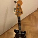 Černá basová kytara s přírodním dřevěným hmatníkem a hlavou. Na hlavě je vidět nápis Fender MUSICMASTER BASS. Nástroj stojí ve stojanu na dřevěné pod…