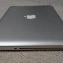 Stříbrný notebook z pohledu zavřeného víka. Uprostřed horní části je bílé logo Apple. Zařízení leží na světlém koberci.