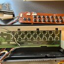 Fotografie zobrazuje vnitřní elektronické součástky hudebního nástroje. V horní části je hnědá deska s řadou kulatých tlačítek a nápisy PANEL B, ASSY…