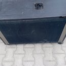 Černý hranatý reproduktorový box s povrchem z textilie. Na horní straně je umístena černá plastová rukojeť a v rozích jsou vidět kovové ochranné rohy…