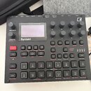 Černý digitální sekvencer a syntetizér s nápisem Syntakt a textem 12 Track Drum Computer & Synthesizer. Zařízení obsahuje displej, několik otočných k…
