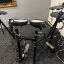 Elektronická bicí souprava na černém kovovém rámu. Na padech a činelech je viditelné logo Roland. Souprava obsahuje dva hlavní pady, dva činely a ped…