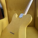 Zadní strana žluté elektrické kytary s tvarem typu Telecaster, opřená o žlutý křeslo. Tělo kytary vykazuje známky opotřebení v podobě škrábanců a odř…