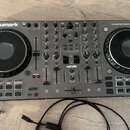 Černý DJ kontrolér s dvěma jog-koly a množstvím potenciometrů, posuvníků a tlačítek. V levém horním rohu je bílý nápis Numark, v pravém horním rohu t…