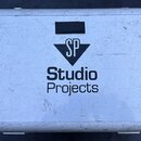 Bílý transportní kufr s kovovými hranami a rohy. Uprostřed víka je černý nápis „Studio Projects“ a nad ním trojúhelníkové logo s písmeny „SP“. Povrch…