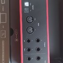 Zadní strana zvukové karty s červeným rámem a černým panelem. V horní části je nápis Focusrite a konektory označené 5V DC a USB. Následují vstupy a v…