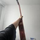 Fotografie zachycuje akustickou kytaru v červenohnaté barvě, kterou drží osoba. Nástroj má hnědý krk a hlavu s ladicími kolíky. Na těle kytary jsou v…