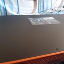 Fotografie zachycuje horní část černého elektronického zařízení s oranžovým okrajem. Na povrchu je nalepený bílý štítek s technickými informacemi, be…