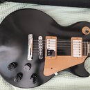 Černá elektrická kytara s kulatým korpusem a béžovým pickguardem. Na těle jsou čtyři černé oválné knofíky, dva humbuckery s bílými kryty a stříbrný m…