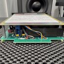 Fotografie zachycuje vnitřní elektroniku zařízení na zelené základní desce s kovovým krytem. Na bílém štítku je text: „Handle the Unit with Care“, „K…