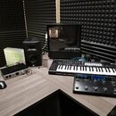 Na hnědém stole v místnosti s akustickými panely je umístěno několik zařízení. Je zde černý klávesový syntezátor s nápisem Novation, černý studiový m…