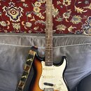 Elektrická kytara typu Stratocaster s barevným přechodem těla od žluté přes oranžovou k hnědé. Na hlavě hmatníku je viditelné logo Fender. Kytara má…