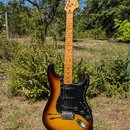 Elektrická kytara s tónem těla v přechodové barvě od černé po žlutou a černým pickguardem. Na hlavě hředu je vidět nápis Squier STRATOCASTER. Kytara…