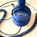 Černá uzavřená sluchátka s pružným spirálovým kabelem a konektorem. Na vnější straně mušle je bílý nápis audio-technica, model ATH-PRO5X a logo značk…