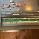 Šedý syntetizátor s nápisy Roland a JUNO-1, který je otevřený a vnitřní elektronika s deskami plošných spojů je viditelná. Na ovládacím panelu je náp…