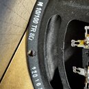 Detail černého kovového rámu reproduktoru s textem v bílé barvě. Jsou zde viditelné nápisy „10/100 TR 8Ω“ a níže řada číslic „220 1630“. Povrch mater…