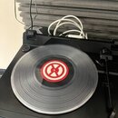 Černý gramofon s umístěnou vinylovou deskou. Uprostřed desky je červenobílý kulatý štítek s grafickým symbolem. V pravé části je vidět tonearm a v po…