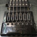 Detailní záběr na černý kovový kobylku kytary s nápisem EVERTUNE. Na fotce jsou vidět šest strun a dvě černé humbuckery s bílými póly. Povrch kobylky…
