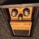 Oranžový kytarový efekt v černé krabici. Na horní části je nápis OverDrive, OD-3 a označení INPUT a OUTPUT se šipkami; v horní části je černé pole s…