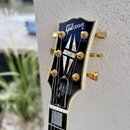 Detail hlavy elektrické kytary v černé barvě s bílým lemováním a zlatými ladícími kolíky. Na horní části je bílý nápis Gibson a v dolní části nápisy…