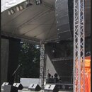 Fotografie zachicuje venkovní koncertní pódium s bílým zastřešením a hliníkovými nosnými konstrukcemi. Vpravo je zavěšený lineární reproduktorový sys…