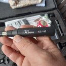 Ruka drží černý cylindrický předmět s kovovým konektorem. Na povrchu je bílý nápis „the ut.bone CC 100“. V pozadí je otevřený černý kufr s vložkou a…