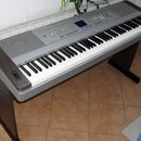 Digitální klavír v šedé barvě s dřevěnými bočními panely a černým stojanem. Na horním panelu je modrý displej, ovládací tlačítka a nápis Yamaha. Povr…