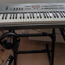 Šedý digitální klavír s bílými a černými klávesami, umístěný na černém stojanu. Na levé straně je nápis KURZWEIL a v pravé horní části označení SP2X.…