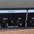 Zadní strana černého elektronického zařízení s bílými popisky. Vlevo je vstup pro napájení s nápisem 12V⎓/1A. Střední sekce OUTPUT obsahuje dva jacky…