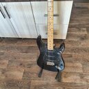 Černá elektrická kytara s tvarem typu Stratocaster a jasně hnědým hnedlem. Kytara má černý pickguard, tři snímače a třicet ladících kolíčků na hlavě.…