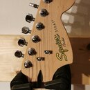 Fotografie zachycuje hlavu elektrické kytary z přírodního světlého dřeva. Na ní je umístěno žluté logo Squier s nápisem by Fender a vedle něj svislý…