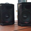 Dva černé reproduktory s kovovými mřížkami v plastových korbelech. Na spodní části obou reproduktorů je červeno-bílé logo s nápisem APOGEE SOUND INC.…