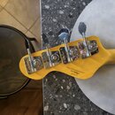 Záběr na hlavu basové kytary z přírodního světlého dřeva s chromovanými kolíky. Na zadní straně hlavy je vidět logo "Fender" a pod ním sériové číslo…