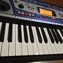 Modrý elektronický klavír značky YAMAHA s bílými a černými klávesami. Na ovládacím panelu jsou vidět různé tlačítka, posuvníky a displej s oranžovým…