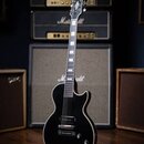 Černá elektrická kytara s tmelem hmatníkem a chromovaným hardwarem, umístěná na stojanu. Na hlavě hmatníku je bílé logo Gretsch a na korpusu pod sním…