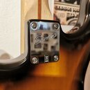 Detail chromovaného krytu hрифu elektrické kytary s nápisem Squier. Kryt je připevněn čtyřmi šrouby a v dolní části obsahuje malý čtvercový symbol s…