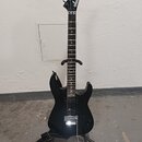 Černá elektrická kytara s špičatou hlavou, na které je bílé logo Jackson. Nástroj má tmavý hmatník a stříbrný kobylku typu Floyd Rose. Kytara je umís…