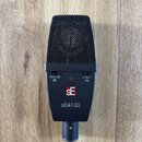 Černý dynamický mikrofon s kovovou mřížkou. Na těle jsou vidět přepínače s nápisy -10 0 -20 dB a 0 Hz, logo SE v červeném čtverci a modelové označení…