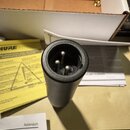Fotografie zachicuje spodní část černého cylindrického zařízení s připojeným konektorem XLR. V pozadí je vidět otevřená kartonová krabice a žlutý pap…