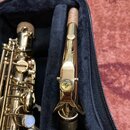 Fotografie zachicuje část zlatého saxofonu a samostatný zlatý příslušený nástroj (neck strap/hnízdový mechanismus) umístěný v černé polstrované kase.…