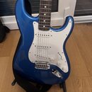 Modrá elektrická kytara typu Stratocaster s bílým pickguardem a hnědým hmatníkem. Nástroj má tři snímače, jeden přepínač a tři bílé ovládací knoflíky…