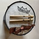 Kruhové buben s bílou membránou, na které je černý nápis REMO USA. Na bubnu leží dva dřevěné paličky s kulatými hlavami, z nichž každá má černé logo…