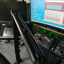 Černý ramenní stativ na mikrofon s kloubovým mechanismem, připevněný k hraně šedého stolu. V pozadí je vidět monitor s rozhraním zvukového softwaru,…
