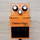 Oranžový kytarový efekt s nápisy Distortion a DS-1. Na horní části jsou tři ovládací knoflíky s popisky TONE, LEVEL a DIST oraz kontrolka CHECK. Ve s…